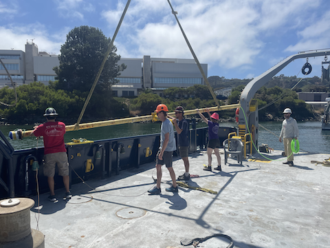 Technicians installing an instrument on a long pole over the side of the ship while it is at the dock